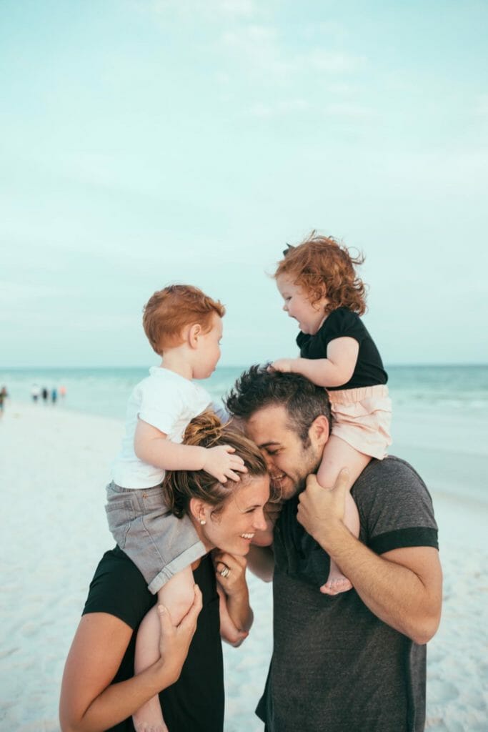 A couple stands on the beach with their read headed twin children on their shoulders.
