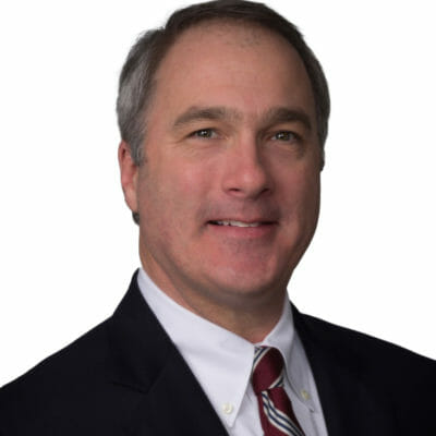 a headshot of a man with grey hair wearing a black suit jacket, white button down shirt and red and white striped tie